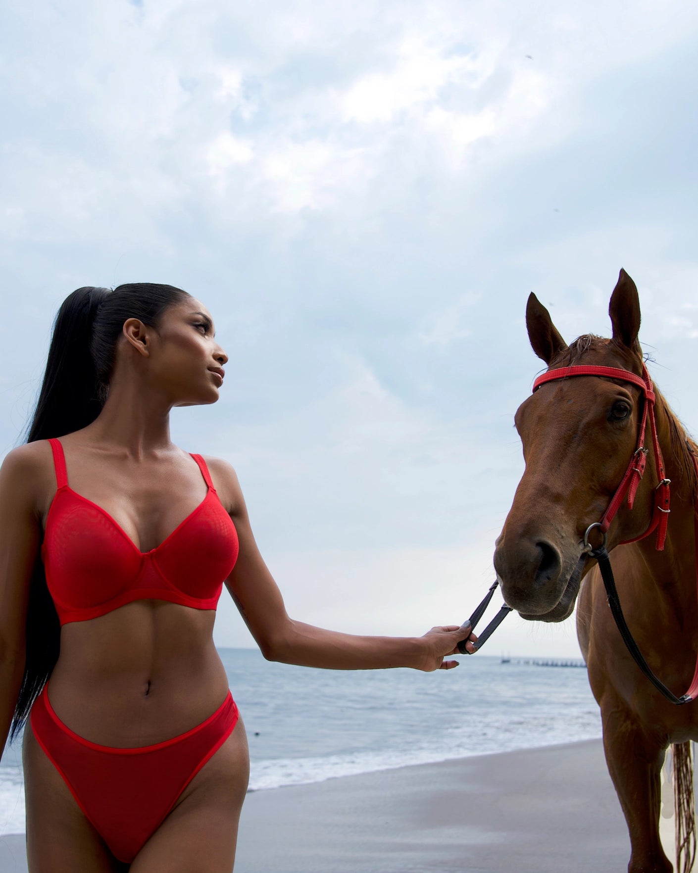 Woman wearing a red sheer mesh underwire bra poses on a beach with a horse