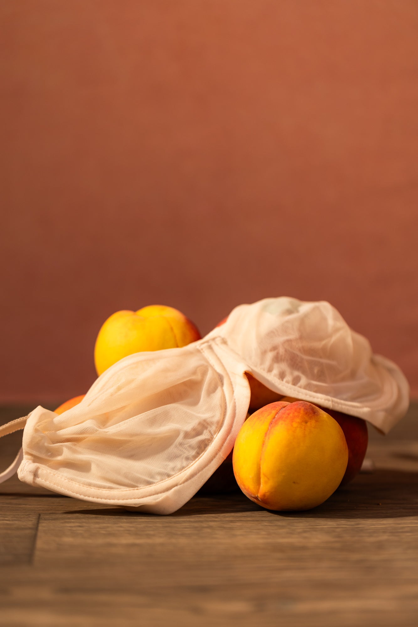 A pale blush beige mesh bralette with matching mesh face mask, styled with fresh yellow-orange peaches on a rustic wooden surface against a warm terracotta background.